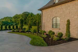 Landscaping around the corner of a stone home with trees in the background