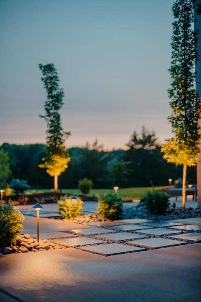 patio of square pavers and pebbles surrounded by bushes, trees, and lighting at sunset