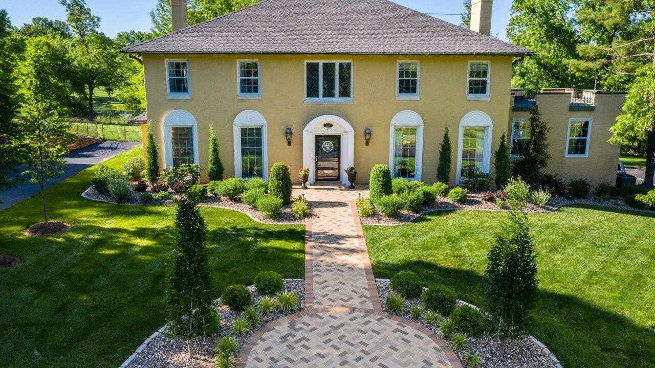 house with paver walkway and landscaped beds with bushes and trees