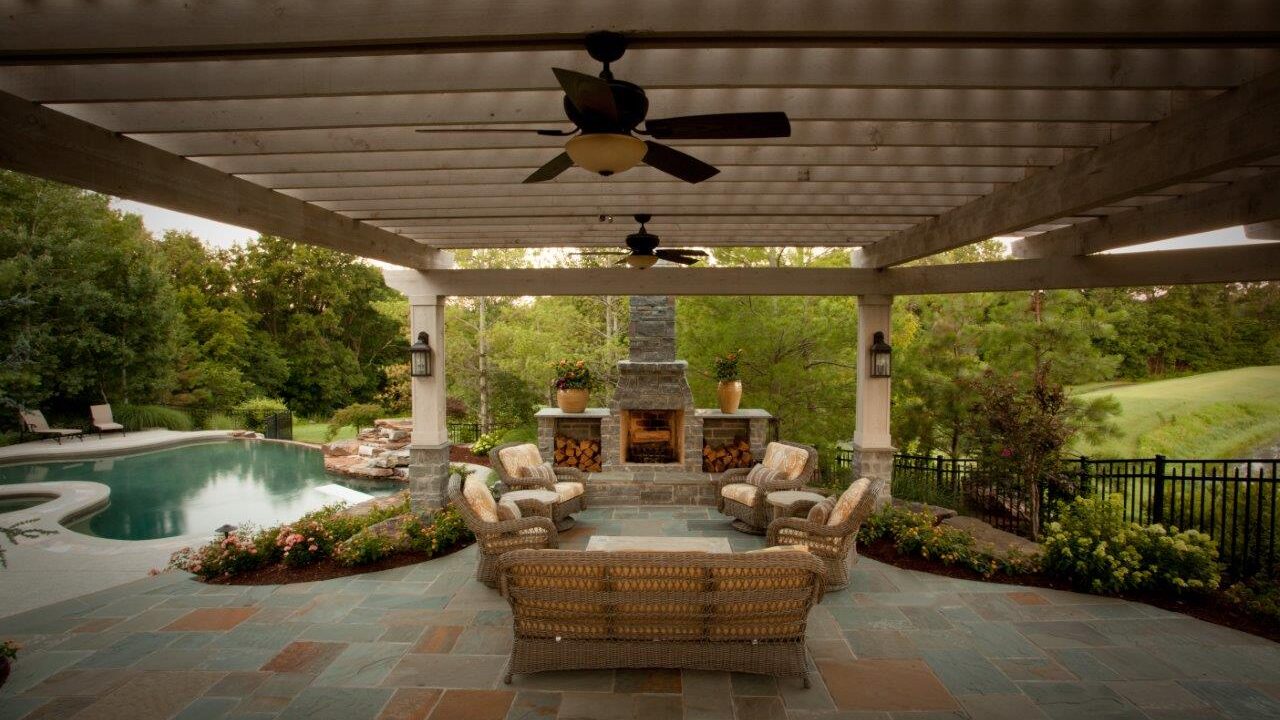 outdoor pergola with fans over seating area with nearby pool and stone fireplace