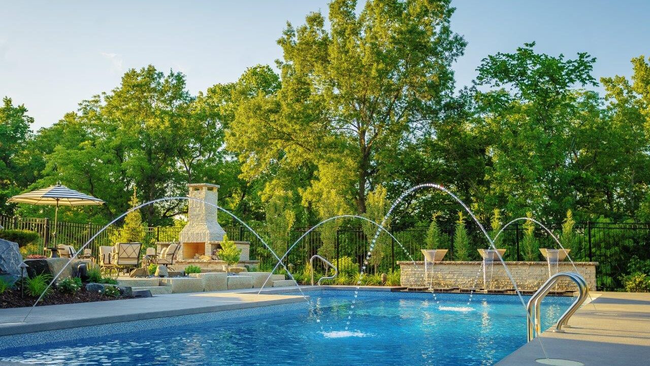 fountains with arching water in a pool with an outdoor fireplace with seating in the background during the day