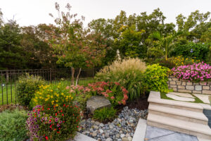 Landscaped yard with shurbs, ornamental grass, flowers, and trees with smooth stones along fence line