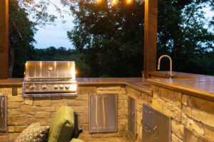 outdoor stone kitchen with sink and grill lit up at night